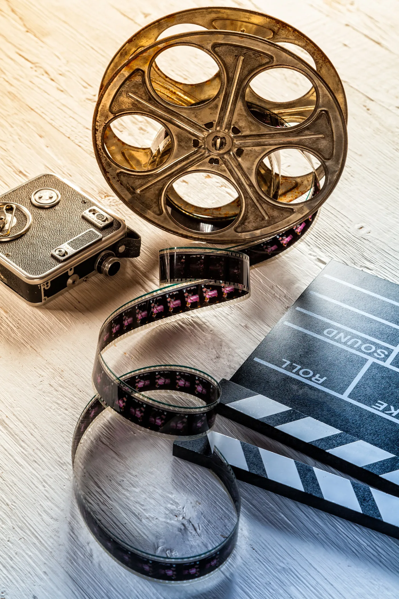 Weathered film reel, vintage movie camera, and open clapperboard on a whitewashed wooden table